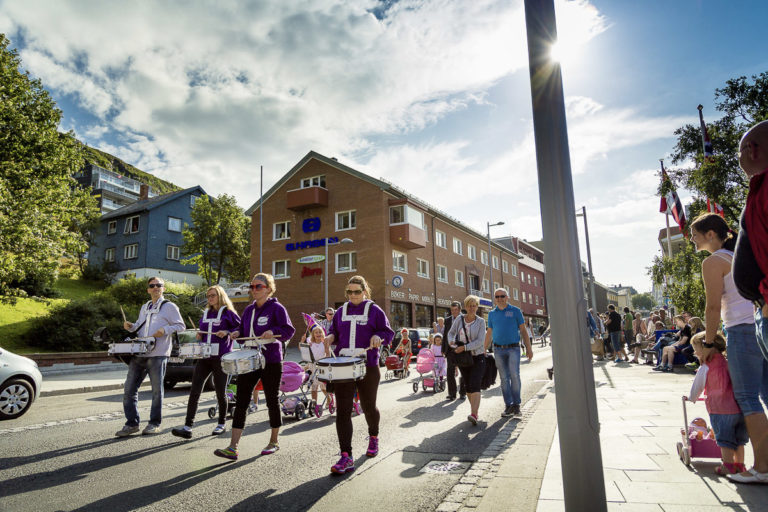 Festlig og fargerik gjenreisningsarkitektur i Hammerfest, med en forretningsgård foran og et typisk gjenreisningshus i blått over. Øverst ligger moderne leiligheter fra Hammerfests oljealder © Ziggi Wantuch